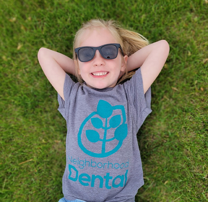 Child with sunglasses laying in grass and wearing Neighborhood Dental Brandon T shirt